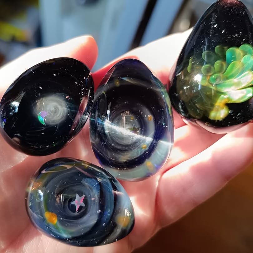 Handmade galaxy teardrop glass pendants held in hand — one holding a green succulent, the others opal planets and shooting stars